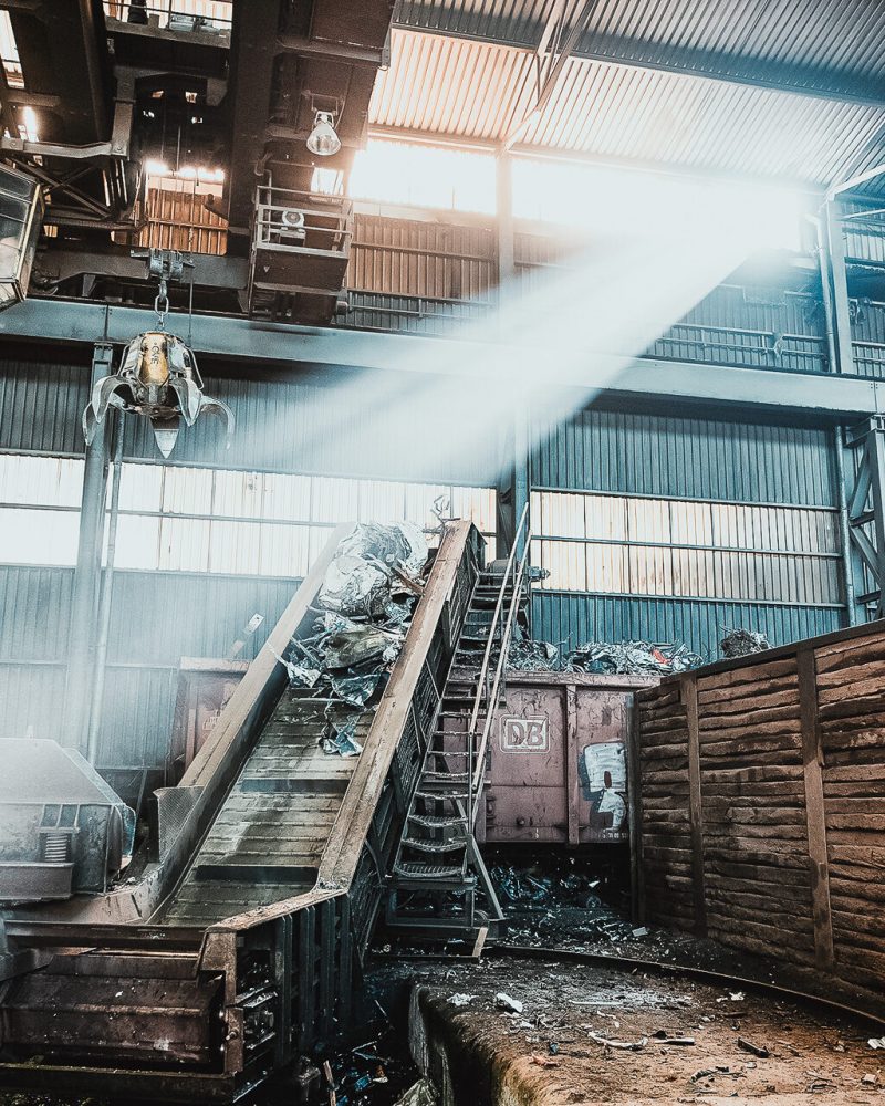 Sonnenlicht str&ouml;mt in eine industrielle Lagerhalle und beleuchtet ein F&ouml;rderband, das mit Metallschrott f&uuml;r das Recycling beladen ist. Holzbarrieren und verstreuter Schrott s&auml;umen den Raum, w&auml;hrend Metalltr&auml;ger und Ger&auml;te &uuml;ber dem Boden h&auml;ngen und den Entsorgungsprozess verdeutlichen. - Altfeld Recycling Ennepetal & Gevelsberg